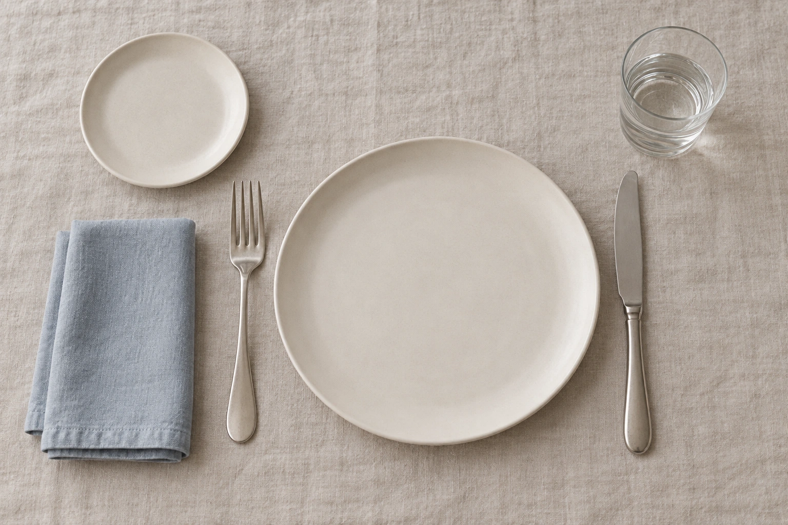 A simple place setting with the fork on the left, knife on the right, bread plate above left and water glass above right.