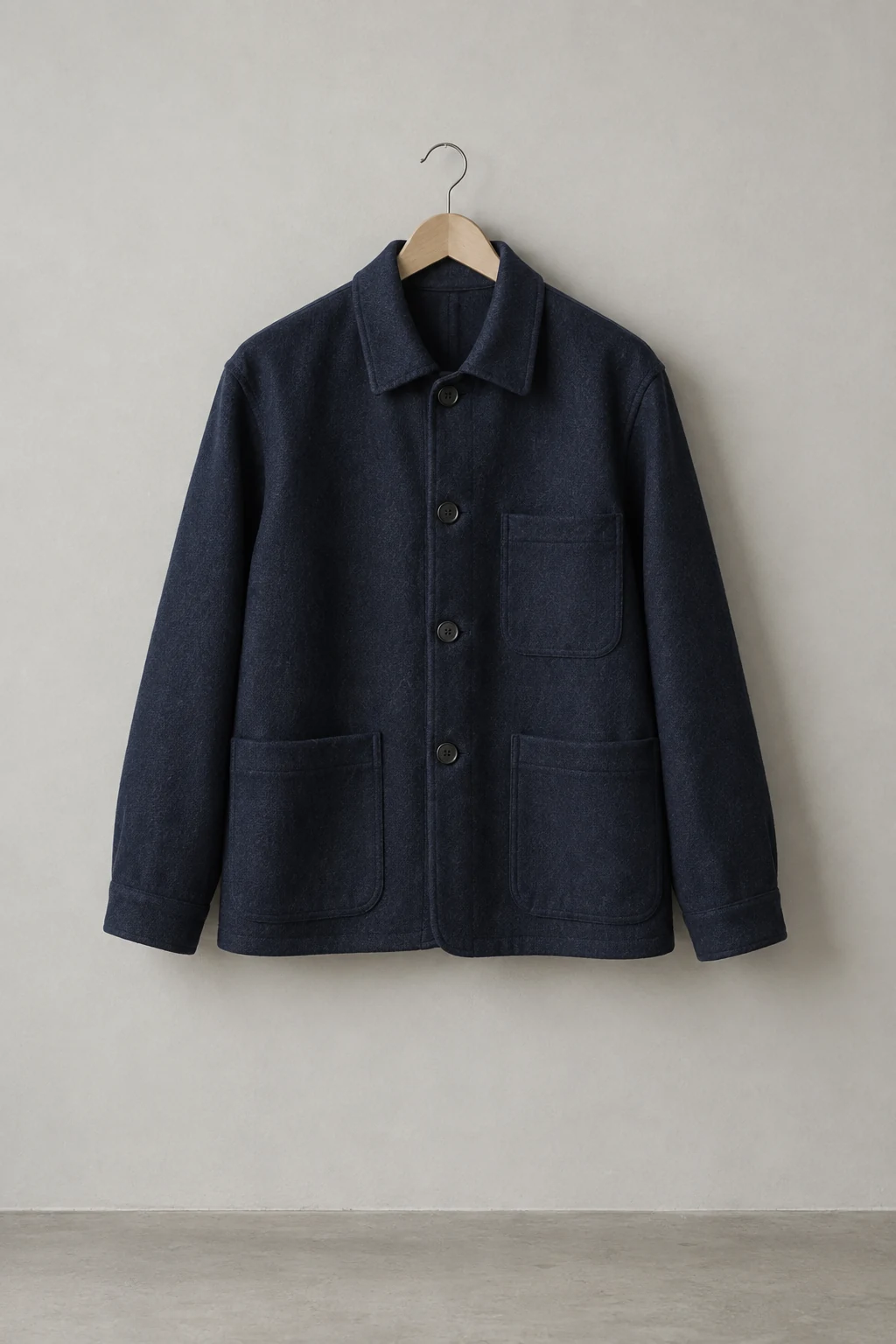 A navy wool chore jacket hangs centered on a wooden hanger against a pale gray studio wall