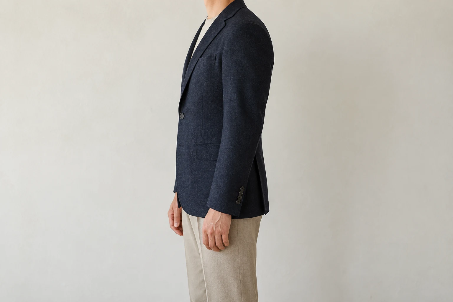 A person shown from the neck down wears a buttoned navy wool blazer in profile against an off white wall.