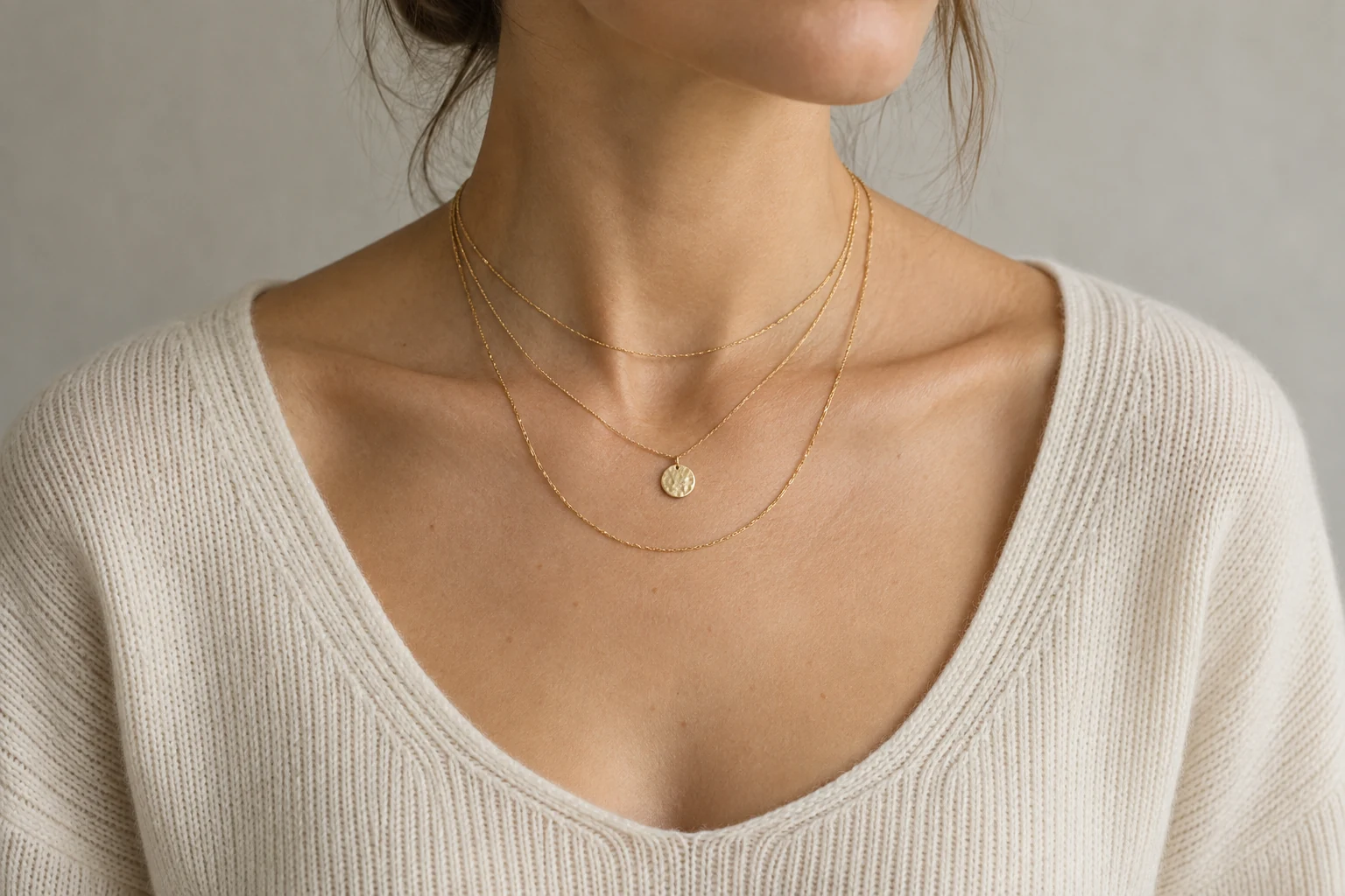 Three fine gold chains rest at distinct lengths over a cream knit neckline with a round pendant on the middle chain.