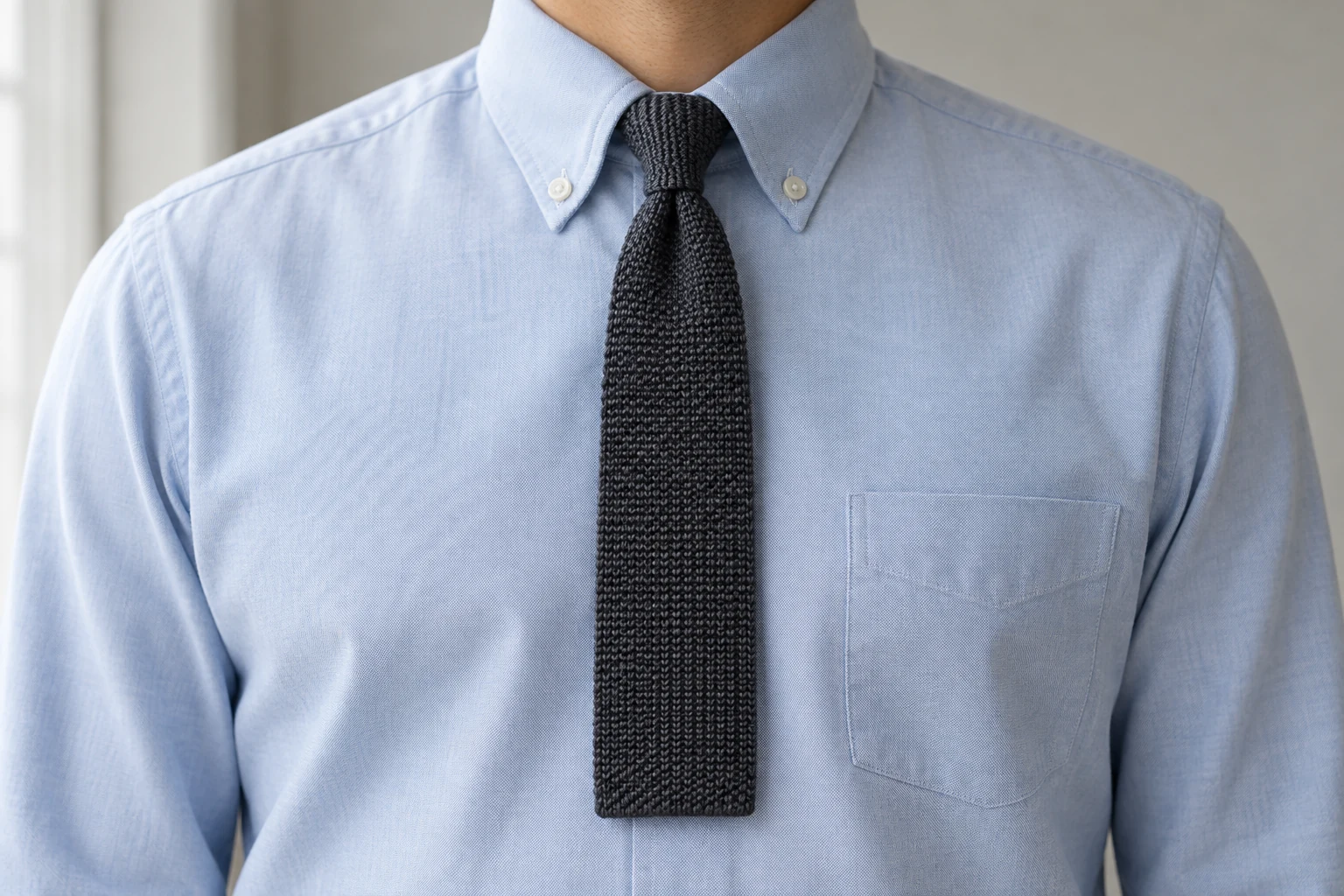 A compact charcoal knit tie knot and square ended blade sit against a pale blue button down shirt