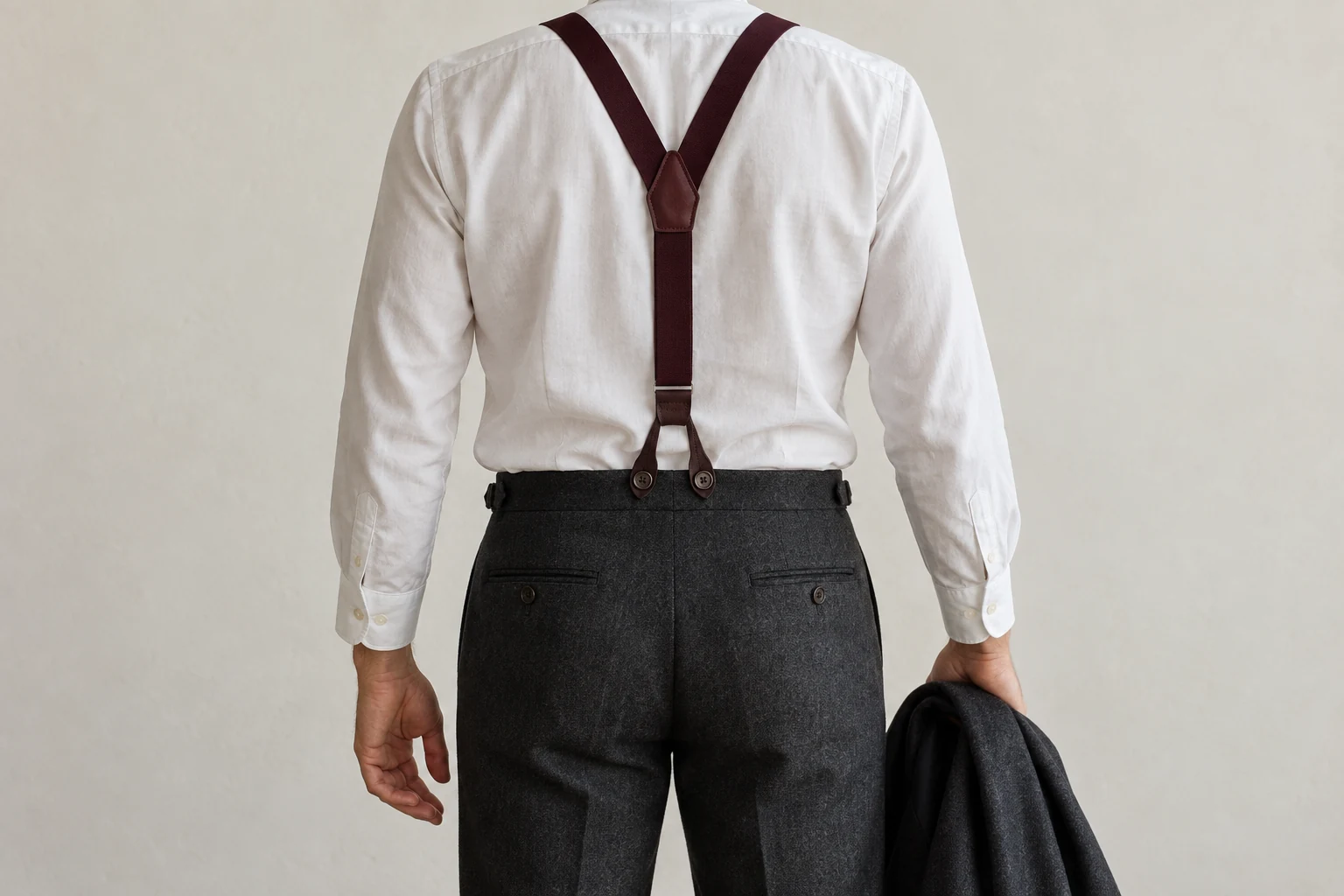 Back view of a man in a white shirt and charcoal trousers wearing burgundy Y back suspenders and holding a jacket.