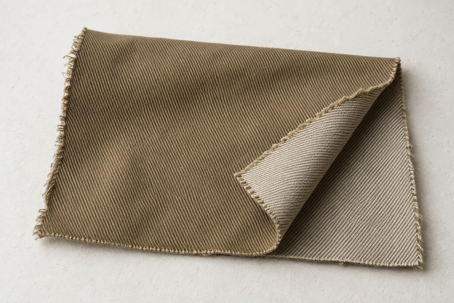 Khaki cotton twill lies folded on an off white surface with diagonal texture and its pale reverse visible.