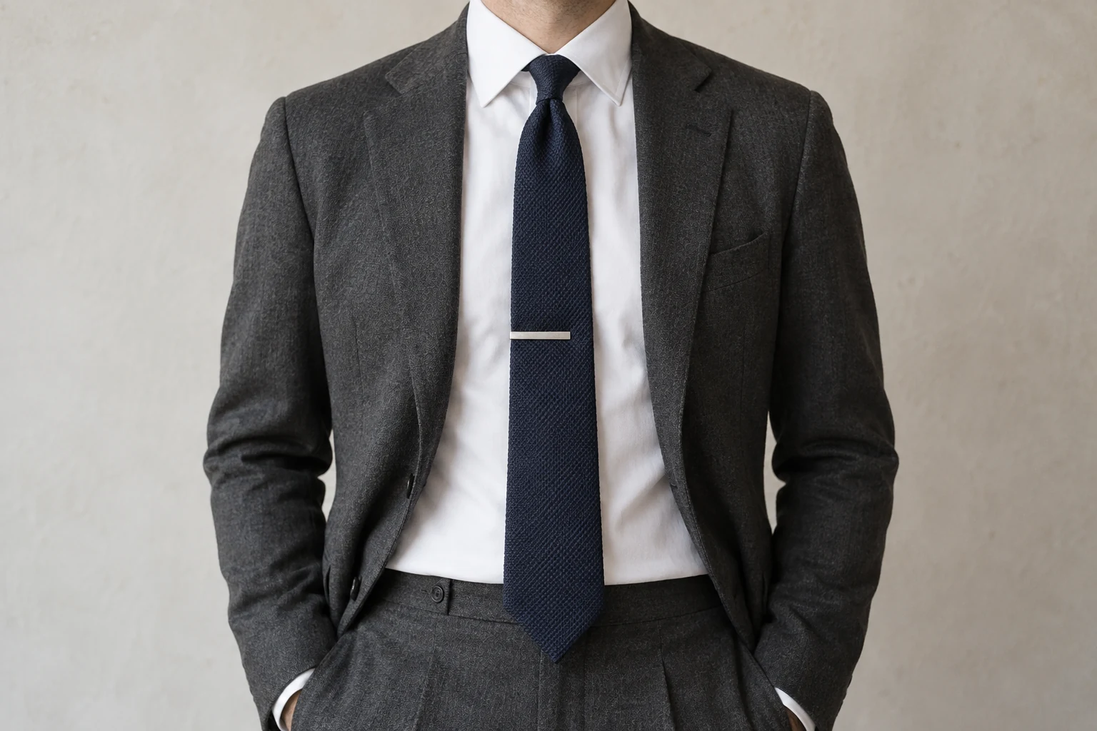 A silver tie bar sits horizontally across a navy grenadine tie framed by a white shirt and charcoal jacket