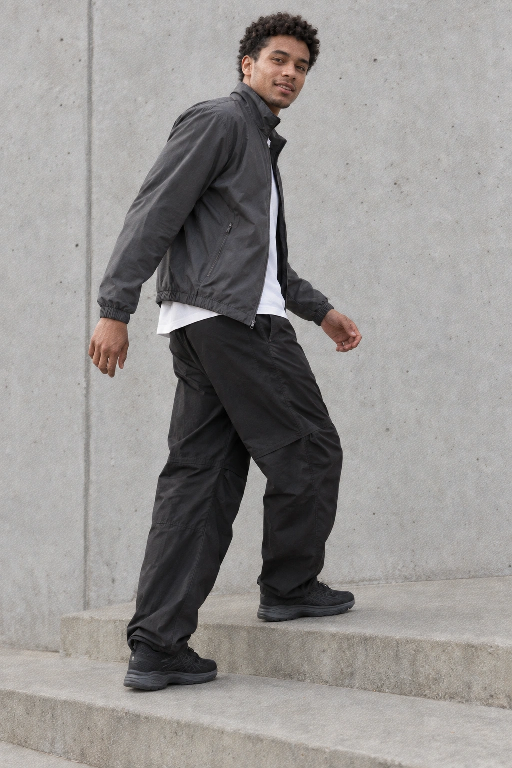 A young man turning toward the camera on a concrete landing, wearing a slate technical jacket and relaxed black trousers.