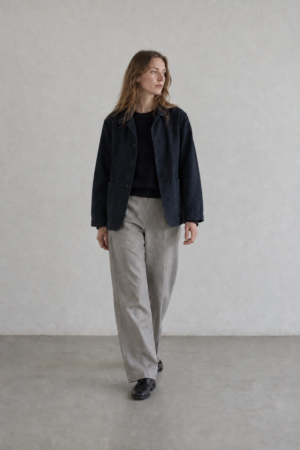 A wider full-body photograph of a long-haired woman stepping across a plain studio floor in a navy cotton jacket and pale trousers.